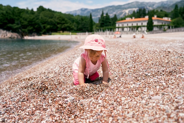 A menina está de quatro na praia perto da água, tendo como pano de fundo a villa Milocer