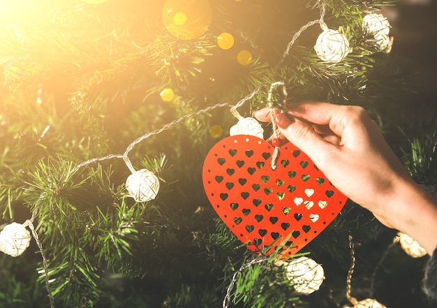 A menina está decorando a árvore de natal com luzes. brinquedo de coração vermelho nas mãos de mulher. conceito de amor e férias. humor de ano novo. estilo hygge. tempo mágico.