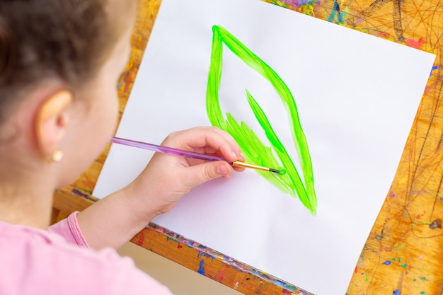 A menina está desenhando folhas verdes com pincel por aquarelas em papel branco em um cavalete. Conceito do Dia da Terra.