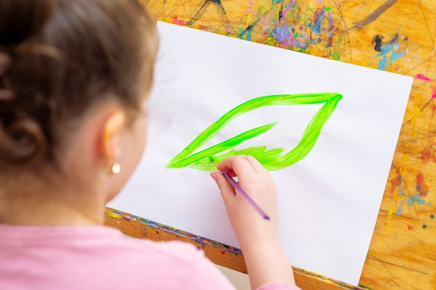 A menina está desenhando folhas verdes com pincel por aquarelas em papel branco em um cavalete. Conceito do Dia da Terra.