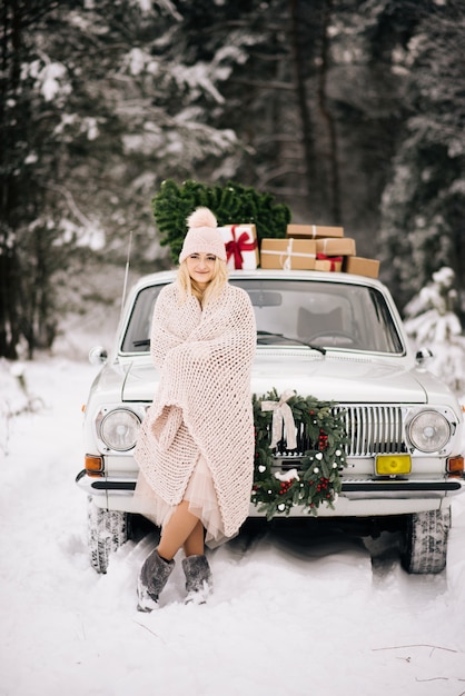 A menina está se preparando para o Natal, é coberta com um cobertor no fundo de um carro retrô, cujo teto é uma árvore de Natal, presentes e uma coroa de flores na floresta de inverno nevado.