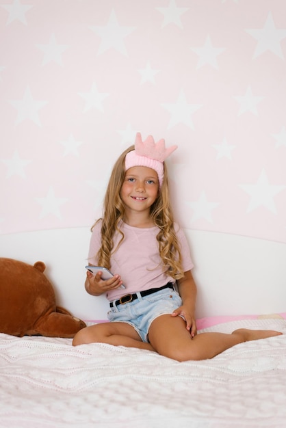 A menina está sentada na cama no quarto rosa segurando o telefone nas mãos e sorrindo