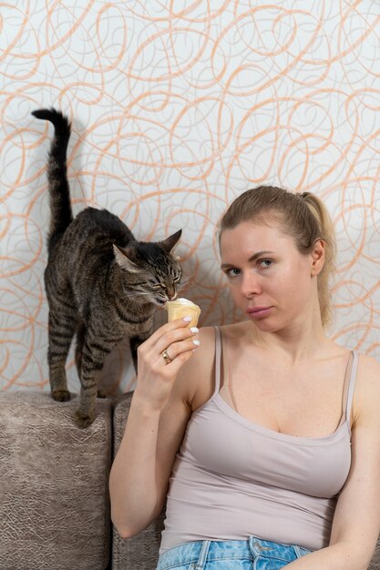 A menina está sentada no sofá segurando um sorvete em um copo de waffle na mão e a gatinha resolve provar esta iguaria