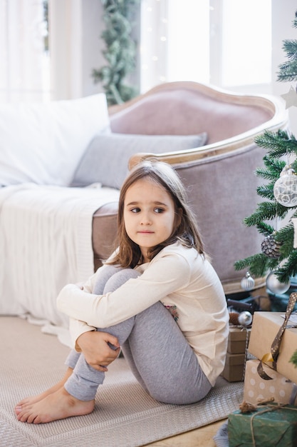 a menina está sentada perto do sofá e decorada com árvore de Natal, a sala está decorada para o Natal