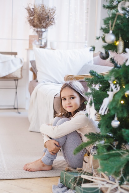 a menina está sentada perto do sofá e decorada com árvore de Natal, a sala está decorada para o Natal