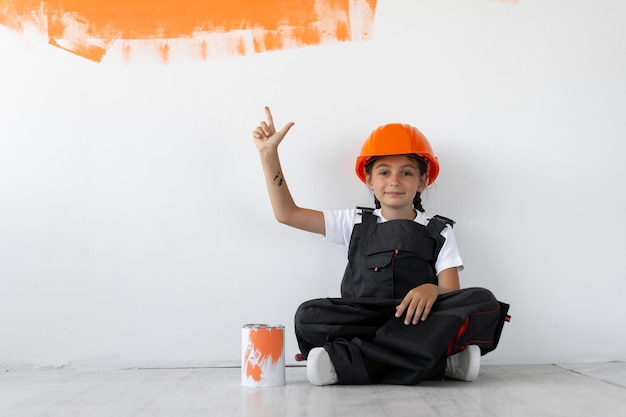 A menina está vestida com um macacão e um capacete protetor laranja na cabeça. sentado no chão olhando para frente. levantou a mão direita com o dedo indicador para cima.