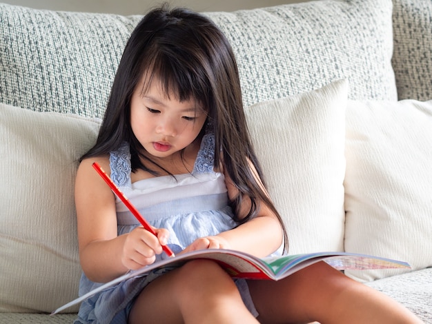 A menina feliz está escrevendo o livro com o lápis vermelho no sofá branco.