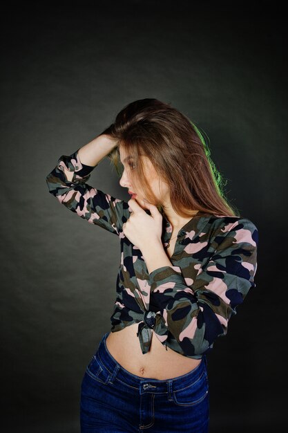 A menina moreno considerável veste a camisa militar e as calças de brim, levantando no estúdio contra o fundo cinzento.