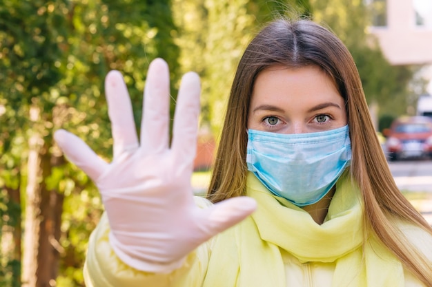 A menina na máscara médica estéril protetora em seu rosto ao ar livre, mostra a palma, mão, não para nenhum sinal. Conceito do coronavirus.