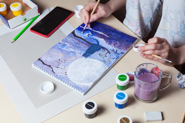 Foto a menina na mesa desenha um gato contra o céu noturno em uma folha branca do álbum em azul na área de trabalho ferramentas frascos aquarela lápis borracha telefone segurando um pincel na mão