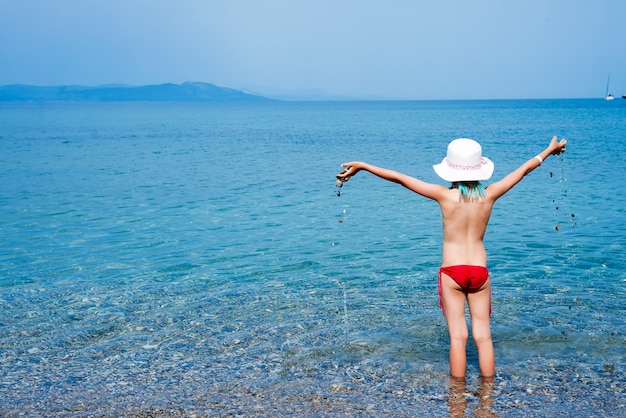 A menina no roupa de banho e no chapéu branco olha o mar e guarda seixos pequenos. Férias de verão no mar Egeu, ilha de Kos, Grécia