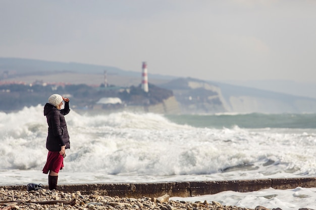 A menina olha para o mar nas ondas de tempestade, ondas de espuma, farol e rochas. a menina está vestida com uma jaqueta preta, chapéu de malha branco, saia longa cor de vinho e botas pretas.