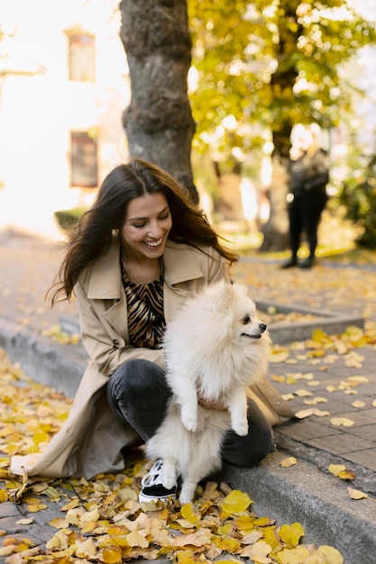 A menina positiva sorridente está segurando um cachorro branco em seus braços sentado folhas amarelas no chão Jovem e seu animal de estimação na rua andando com um animal de estimação