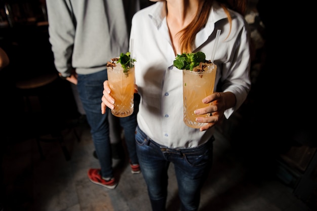 A menina prende dois cocktail alcoólicos alaranjados com gelo, hortelã e canela