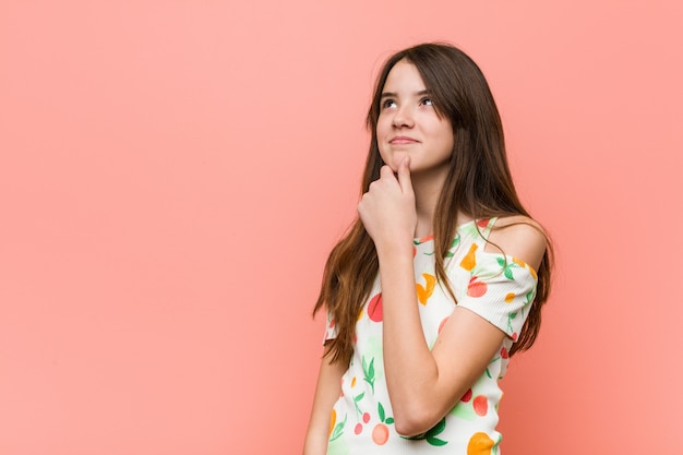 A menina que veste um verão veste-se contra uma parede vermelha que olha lateralmente com expressão duvidosa e cética.