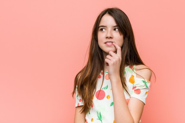 A menina que veste um verão veste-se contra uma parede vermelha que olha lateralmente com expressão duvidosa e cética.