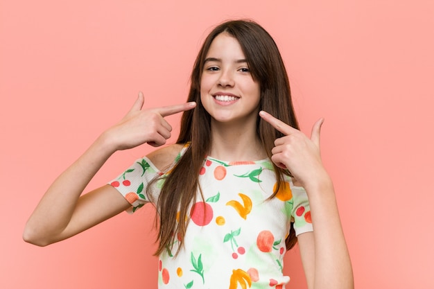 A menina que veste uma roupa do verão contra uma parede vermelha sorri, apontando os dedos na boca.