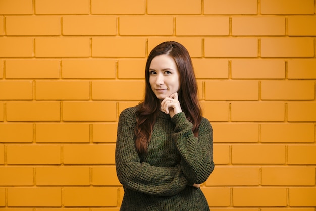 A menina ruiva bonita que veste vidros e uma camisola verde está sorrindo contra um fundo amarelo da parede de tijolo.