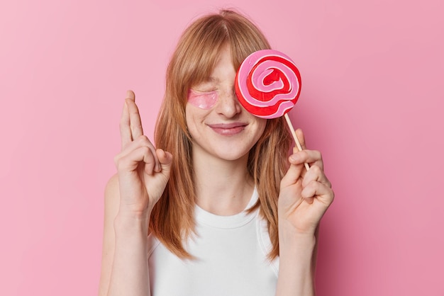 A menina ruiva esperançosa positiva cobre o olho com pirulito doce mantém os dedos cruzados acredita em boa sorte weas camiseta branca casual posa sobre fundo rosa Adolescente ruiva adorável com doces