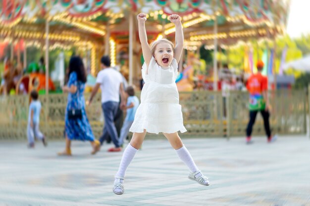 A menina salta e sorri de felicidade em um parque de diversões no verão