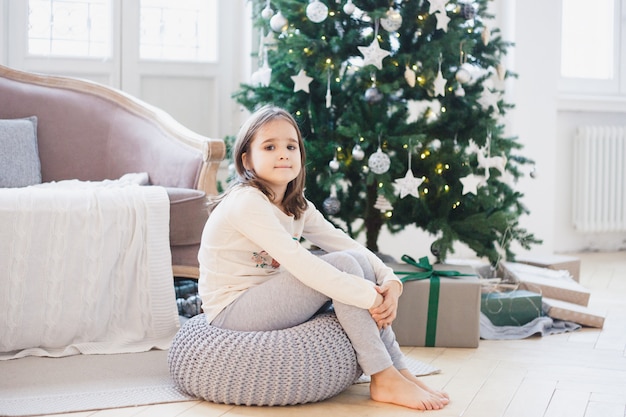 a menina senta-se no otomano ao lado da árvore de natal, a sala está decorada para o natal