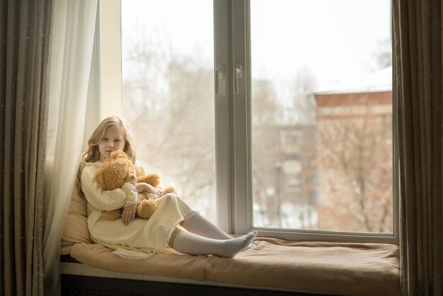 A menina senta-se no parapeito da janela com um brinquedo e olha pela janela