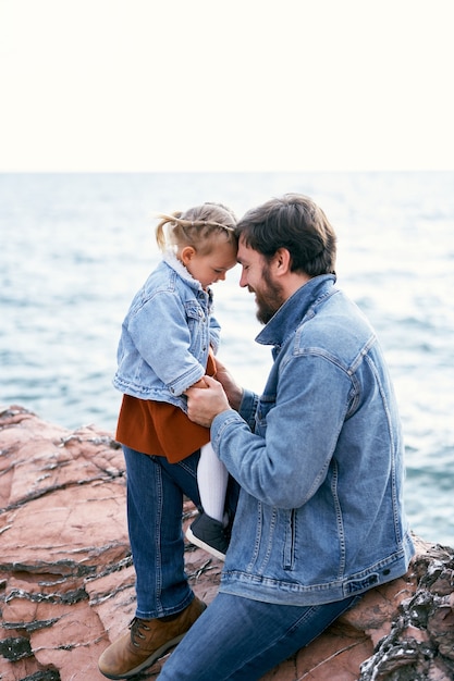 A menina senta-se nos pés do papai, curvando a testa na testa dele, o papai senta-se nas pedras contra o