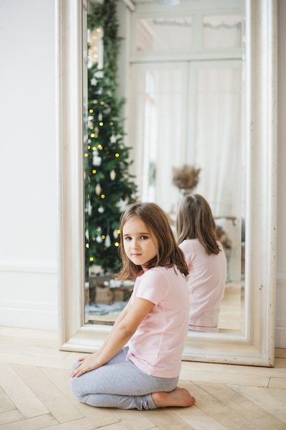 a menina senta-se perto do espelho, o espelho reflete a árvore de Natal e as luzes, o interior é decorado, natal, esperando o feriado