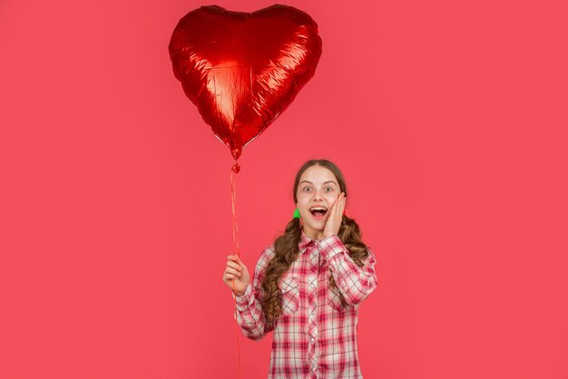 A menina surpreendida segura o balão do coração do amor no fundo vermelho