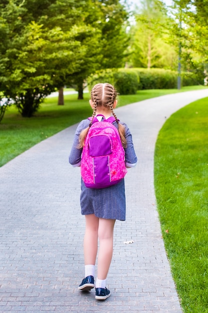 Foto a menina vai estudar. vista traseira no parque. de uniforme e com mochila escolar. escola primária, primeira série