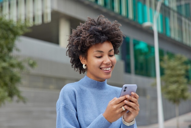 A metade do comprimento da foto de uma mulher feliz e positiva em poses de gola alta azul na urbanidade com telefone celular explora o novo aplicativo de downloads da cidade e conversa por bate-papo. tecnologia de pessoas e tempo livre.