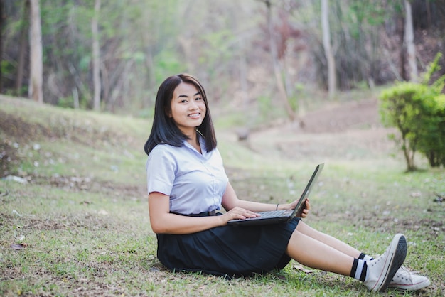 A moça asiática na farda da escola está usando o portátil para a educação e a comunicação no campo de Tailândia.