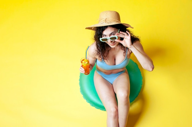 A moça no maiô azul e no chapéu de palha com um círculo de natação inflável senta-se na praia