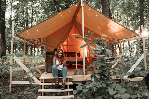 A moça senta ao lado da barraca e bate papo com streaming de mensagens no smartphone, postando, curtindo nas redes sociais. Viaje para fora da cidade na floresta. Acampamento. Férias de baixo orçamento em barraca glamorosa