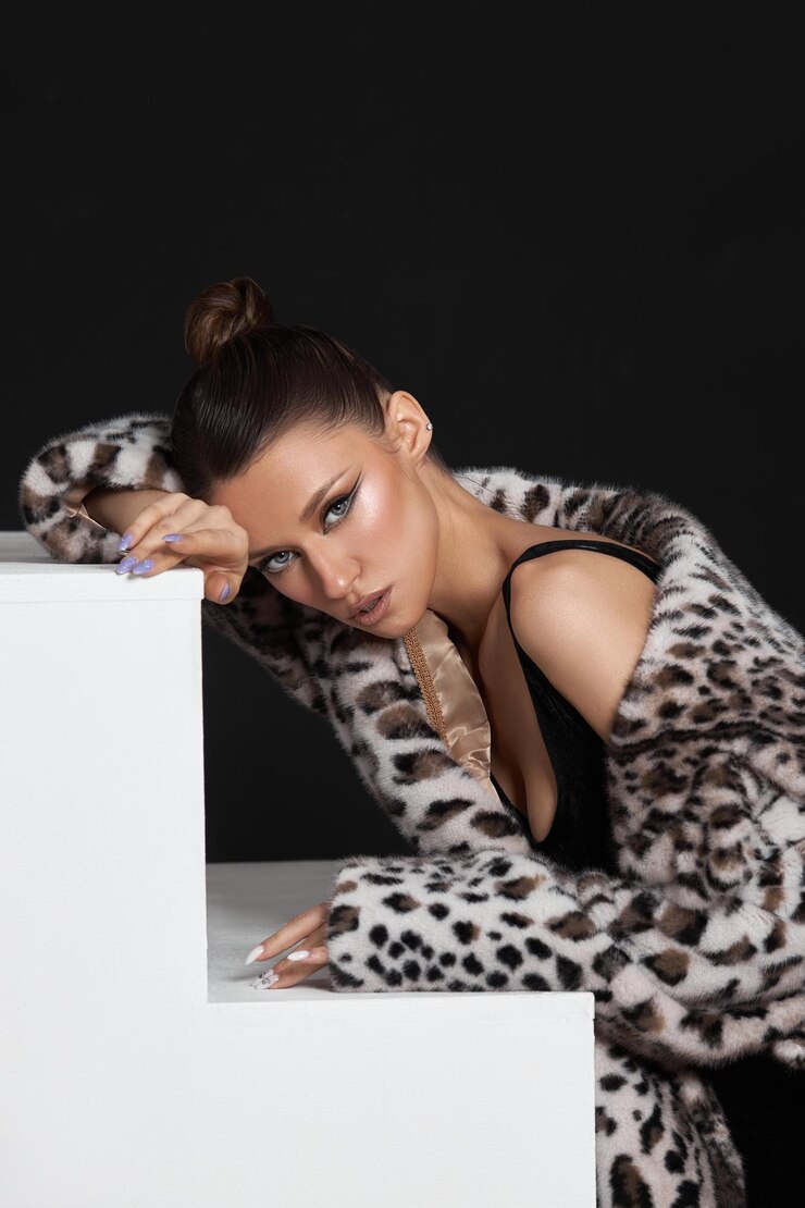 A modelo de beleza se inclina graciosamente sobre um pedestal branco exibindo seu elegante casaco de impressão de leopardo e maquiagem impressionante em um ambiente de estúdio minimalista. O contraste destaca sua pose e expressão.