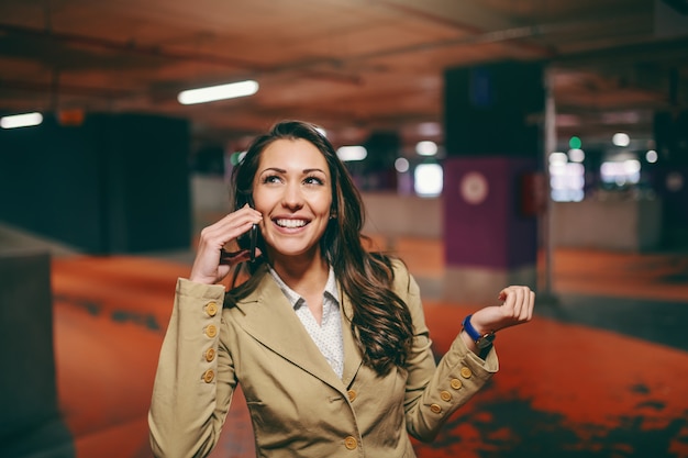 A morena caucasiano de sorriso vestiu elegante usando o telefone esperto e olhando acima no estacionamento subterrâneo.