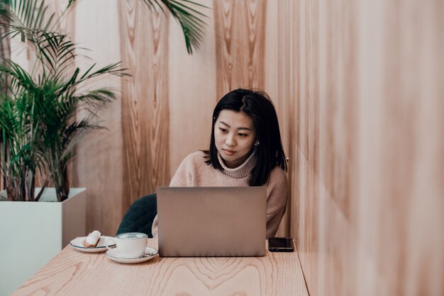 A mulher adulta bonita joga em um laptop em uma cafeteria. Mulher asiática, trabalhando em um café em um laptop. Conceito freelance