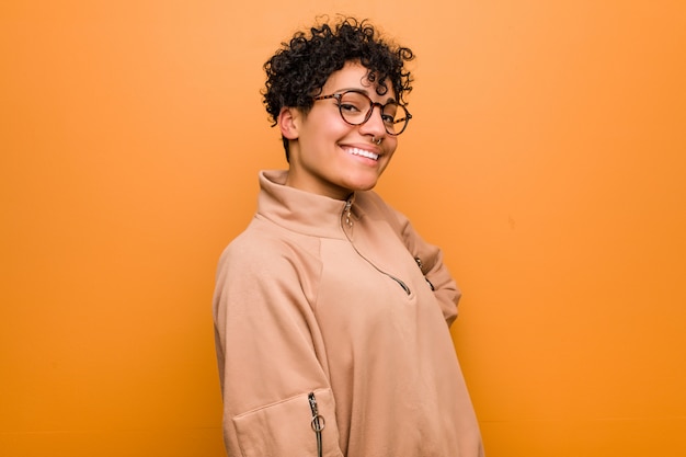 A mulher afro-americano misturada nova contra uma parede marrom olha de lado o sorriso, alegre e agradável.