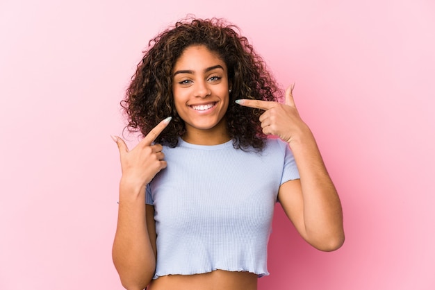 A mulher afro-americano nova contra uma parede cor-de-rosa sorri, apontando os dedos na boca.