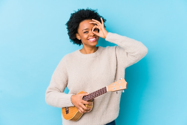 A mulher afro-americano nova que joga o ukelele isolou entusiasmado mantendo o gesto aprovado no olho.