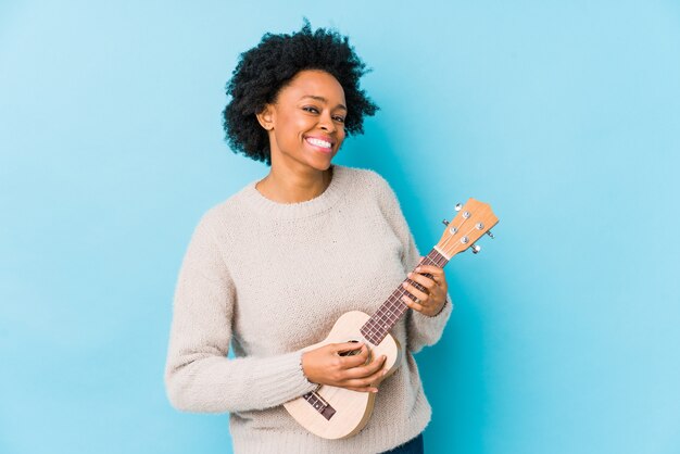 A mulher afro-americano nova que joga o ukelele isolou feliz, sorrindo e alegre.