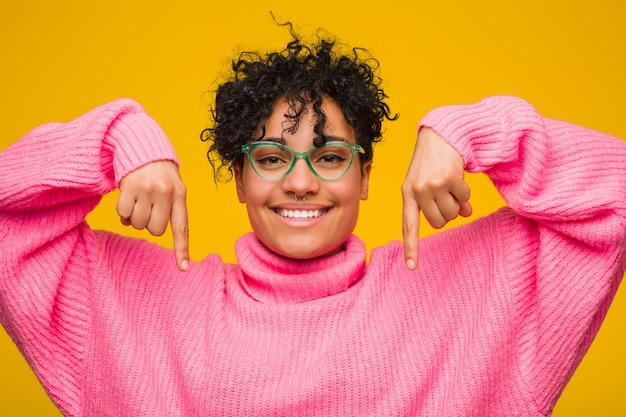 A mulher afro-americano nova que veste uma camisola cor-de-rosa aponta para baixo com os dedos, sentimento positivo.