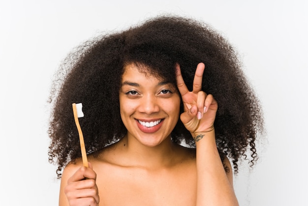 A mulher afro nova que mantem uma escova de dentes isolada mostrando o sinal da vitória e sorrindo amplamente.