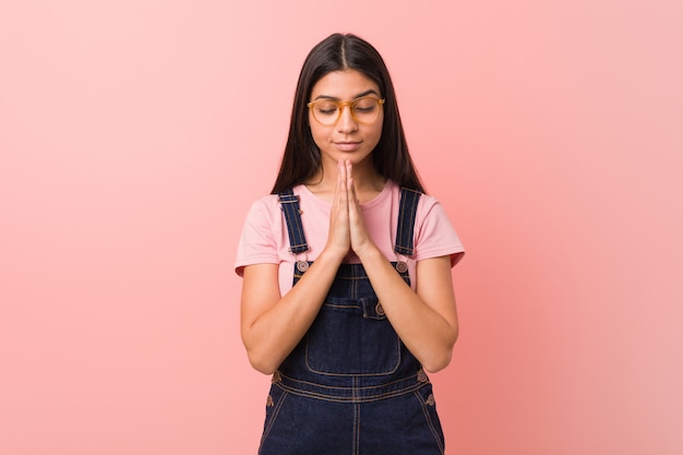 A mulher árabe bonita nova que veste um dungaree das calças de brim que guarda as mãos reza perto da boca, sente-se confiante.