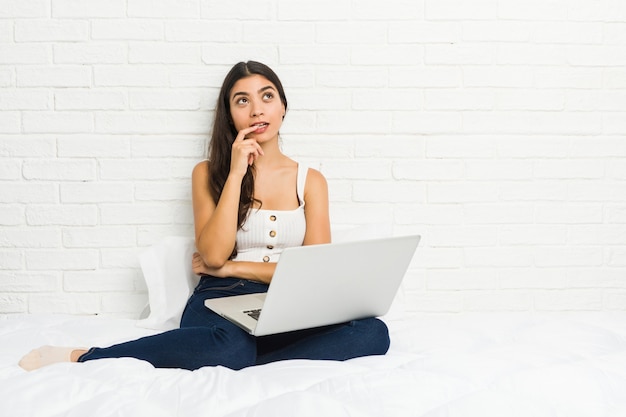 A mulher árabe nova que trabalha com seu portátil na cama relaxou o pensamento sobre algo que olha um espaço da cópia.