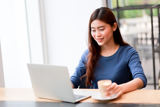 A mulher asiática que usa o computador portátil e bebe o café trabalha do conceito home