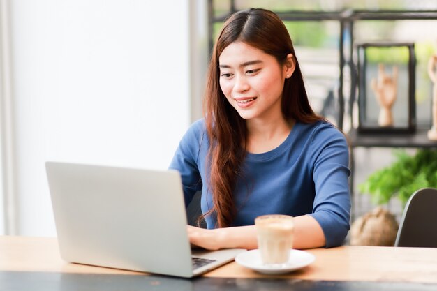 A mulher asiática que usa o computador portátil e bebe o café trabalha do conceito home