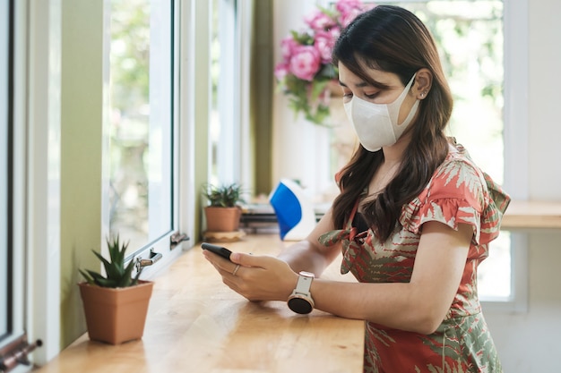 A mulher asiática que veste a máscara protetora e usando o smartphone no restaurante, protege a inflexão de coronavírus. distanciamento social, nova normalidade e vida após pandemia de covid-19