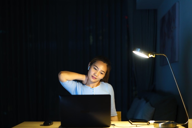 A mulher asiática se estica preguiçosamente enquanto trabalha longas horas na frente de um computador tarde da noite na sala de estar em casa.