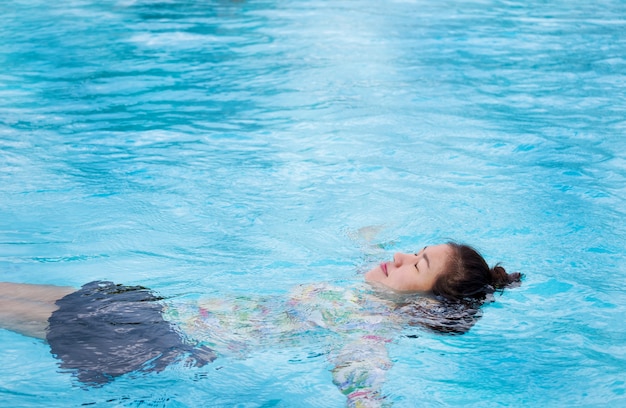 A mulher asiática veste um maiô flutuando em uma piscina ao ar livre com água azul clara nas férias de verão.